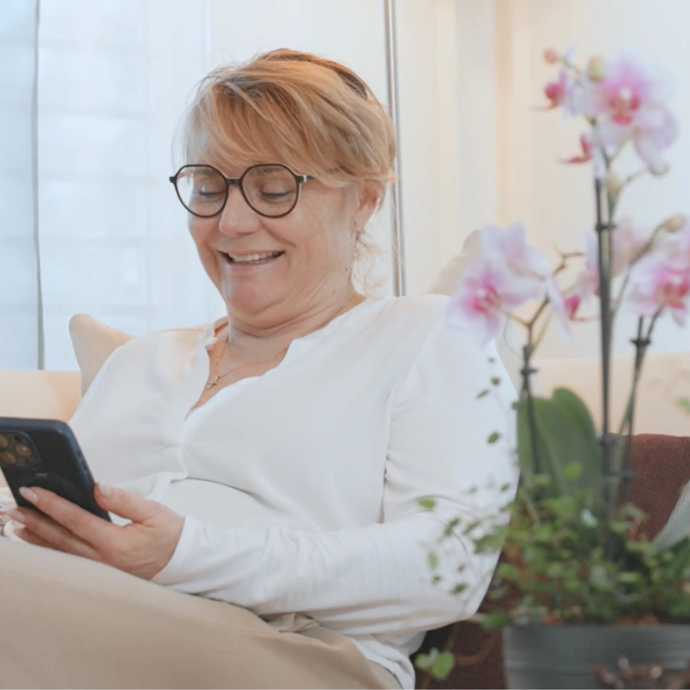 Frau mit Brille sitzt lächelnd am Smartphone mit Blumen im Hintergrund