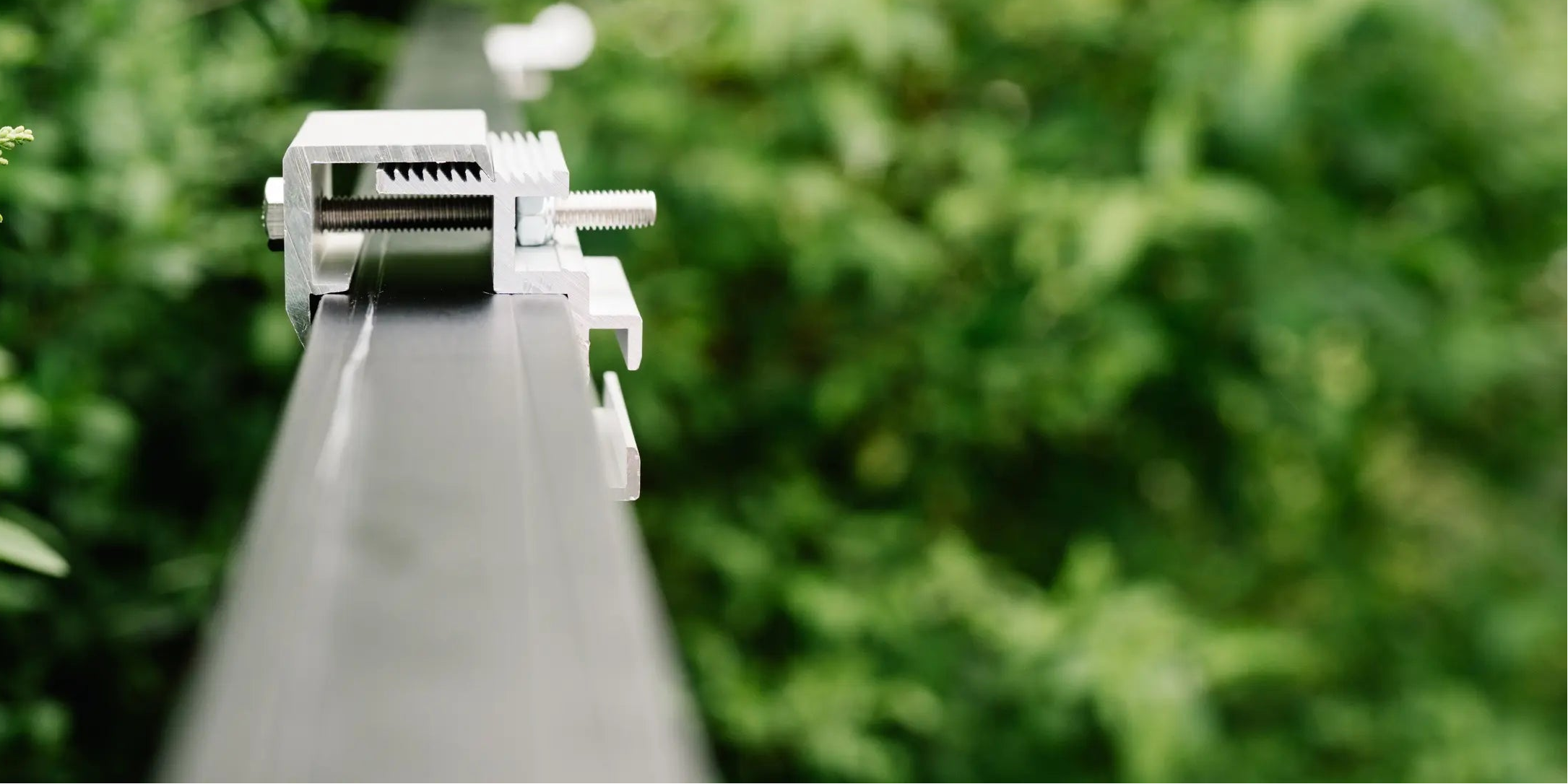 Detailaufnahme einer Metallklemme zur Befestigung eines Solarmoduls auf einer Schiene, mit unscharfem, grünem Hintergrund aus Pflanzen.